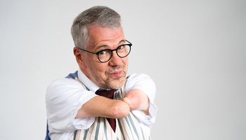 Rainer Schmidt hat die verkürzten Arme ohne Hände zusammengelegt und schaut verschmitzt durch eine Brille den Betrachter an.