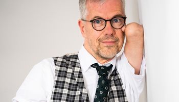 Rainer Schmidt lehnt an einer Wand und hat den verkürzten Arm ohne Hand mit einem Daumen an die Wange gelegt. Er trägt eine Brille.